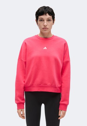 Personne aux cheveux courts et foncés portant un sweat Adidas rose vif et un pantalon noir, debout devant un fond clair uni.