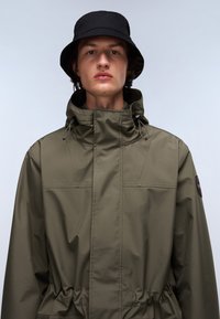 Olivengrüne isolierte Jacke mit hohem Kragen, verstellbarem Kordelzug in der Taille und gummiertem Logo-Patch. Kombiniert mit einem schwarzen Bucket Hat.