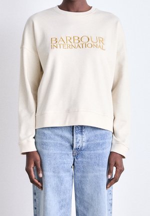 Osoba ubrana w kremową bluzę Barbour International i jasnoniebieskie dżinsy z wysokim stanem, stojąca na tle jednolitego jasnego tła.