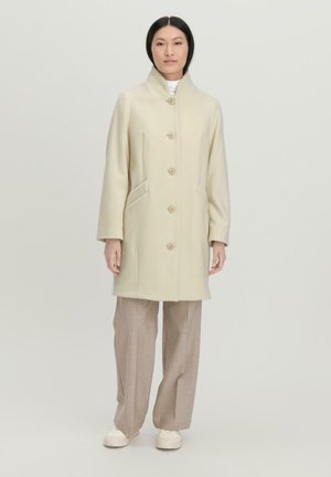 Beige Wollmantel mit hohem Kragen, fünf Knöpfen und zwei Seitentaschen, kombiniert mit hellbeigen weit geschnittenen Hosen und weißen Sneakern.