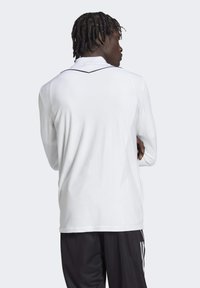 Witte sporttrui met lange mouwen van een gestructureerde stof, met een zwarte accent langs de kraag en schoudernaden, gedragen met zwarte broek.