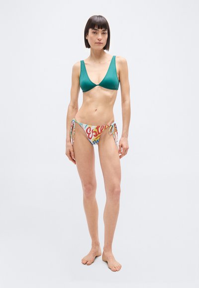 Λεπτή γυναίκα με κοντά σκουρόχρωμα μαλλιά, που φοράει τοπ μπικίνι σε τιρκουάζ χρώμα και πολύχρωμο μαγιό με κορδόνια στο πλάι, στέκεται ξυπόλητη σε λευκό φόντο.