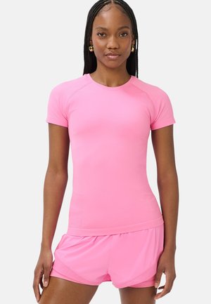 Vrouw met lang gevlochten haar, draagt gouden creolen, een roze atletisch shirt met korte mouwen en bijpassende roze hardloopshorts.