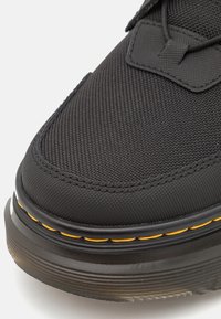 Zapato de textil negro con superficie texturizada, puntera redonda y costuras amarillas contrastantes a lo largo de la suela, con un diseño duradero.