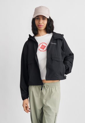 Kvinde iført en sort Columbia jakke, hvid Columbia Sportswear t-shirt, lysegrønne bukser og en lyserød kasket, stående med den ene hånd i lommen.