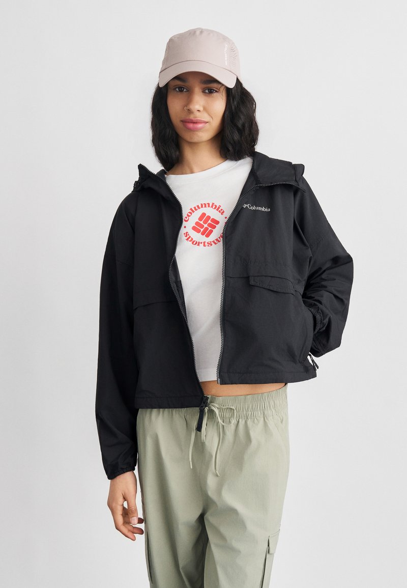 Γυναίκα που φοράει μαύρο μπουφάν Columbia, λευκό t-shirt Columbia Sportswear, ανοιχτοπράσινο παντελόνι και ανοιχτό ροζ καπέλο, στέκεται με το ένα χέρι στην τσέπη.