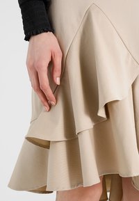 Beige lagerkjol med volanger, tillverkad av slätt tyg. Har en utsvängd fåll och subtila texturdetaljer. Hand som vilar på tyget.