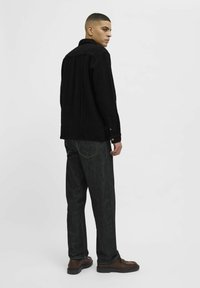 Chaqueta de pana negra con cuello, puños abotonados y canalones verticales visibles, combinada con jeans de mezclilla oscura y zapatos marrones.
