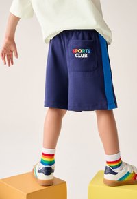 Tamsiai mėlynos šortai su mėlynu šoniniu dryžiu, nugarinėje kišenėje siuvinėtas įvairiaspalvis užrašas „SPORTS CLUB“. Derinamos su vaivorykštės spalvų dryžuotomis kojinėmis.