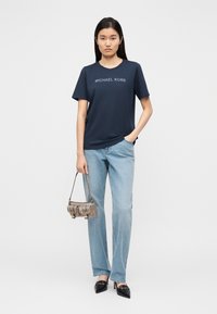 T-shirt de algodão azul-marinho com o logótipo "MICHAEL KORS", combinado com jeans de ganga azul clara e uma mala prateada texturizada. Sapatos de salto alto pretos com bico fino.