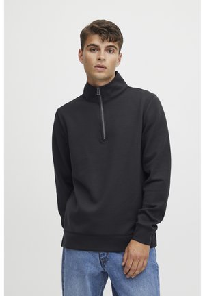Svart zip-genser med høy krage, myk stoff, lange ermer og en avslappet passform. Brukt over lyseblå jeans.
