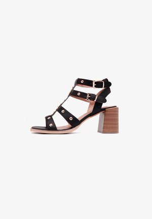 Sandale noire à talons avec brides, ornée de clous dorés, fermetures à double boucle, et talon bloc en bois sur fond blanc.