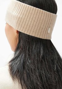 Ribbad beige huvudband gjort av mjukt tyg, med en logotagg och utformat för att täcka öronen, med en snug passform runt huvudet.