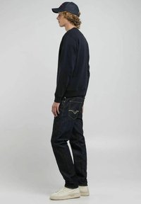 Felpa blu scuro, jeans in denim scuro con dettagli nelle tasche posteriori, sneakers bianche e un cappellino blu scuro, visti di lato.