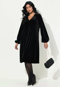 Robe en velours noir avec de longues manches bouffantes, un décolleté en V et une garniture florale. Associée à des collants transparents et un sac à main noir texturé.