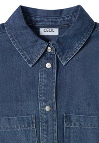 Džínová košile s límečkem, modrý materiál, stříbrné tlakové knoflíky a dvě přední kapsy. Má hladkou texturu a prošívané detaily.