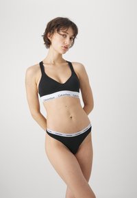 Črn bombažni modrček z V-izrezom in širokim elastičnim trakom, ki vsebuje logotip "Calvin Klein"; ujemajoči se črni bikini z podobnim logotipom na pasu.