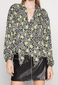 Blusa a maniche lunghe con un motivo floreale in nero, bianco e verde neon, caratterizzata da un profondo scollo a V e dettagli sui polsini arricciati, abbinata a una gonna di pelle nera.