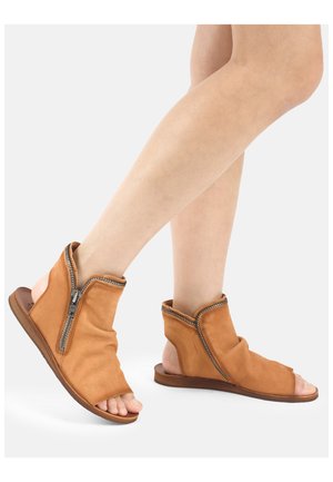 Füße, die beige Leder-Sandalen mit offenen Zehen, seitlichen Reißverschlüssen und offenen Fersen tragen, vor einem schlichten hellen Hintergrund gezeigt.