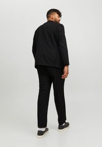 Čierny oblek na mieru so sakom s jedným gombíkom, slim-fit nohavicami a textúrovanou látkou. Čierne topánky s bielymi akcentmi dopĺňajú vzhľad.