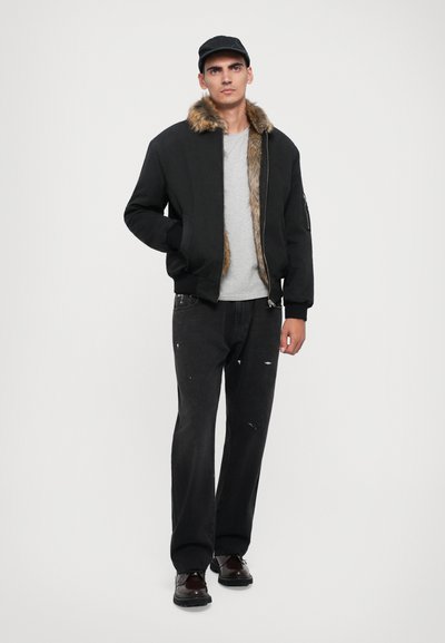 Svart bomberjacka med pälskrage, grå t-shirt, slitna svarta jeans och bruna skor; stående figur mot en ljus bakgrund.