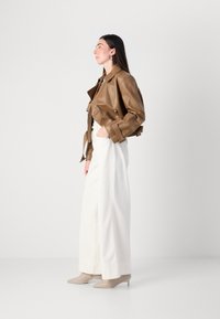 Vrouw in profiel, gekleed in een bruin leren cropped jack, witte wijde broek en beige hoge hakken, met een minimalistische achtergrond.