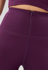 Dopasowane fioletowe legginsy o gładkiej strukturze, z zamkiem błyskawicznym z tyłu oraz wyraźnym przeszyciem wzdłuż szwów.