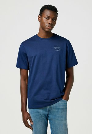 T donkerblauwe katoenen t-shirt met korte mouwen en een ronde hals. Bevat witte tekst op de linkerkant van de borst. Gecombineerd met lichtblauwe jeans.
