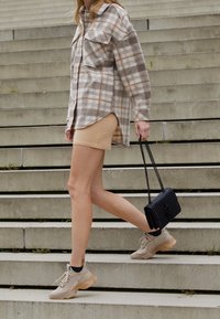 Osoba w beżowych trampkach, brązowej spódnicy i koszuli w kratę wchodzi po betonowych schodach, trzymając małą czarną torebkę z łańcuszkiem.