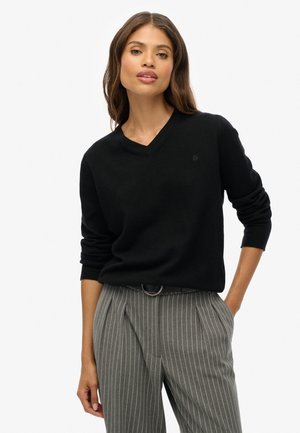 Schwarzer V-Ausschnitt-Pullover aus weichem Stoff, mit langen Ärmeln und taillierter Form, kombiniert mit grauen Pinstripe-Hosen und einem schwarzen Gürtel.