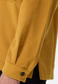Camicia di lana color senape con finitura testurizzata, dotata di polsini con bottone e spacchi laterali per facilitare i movimenti.