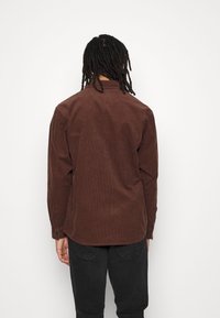 Camicia in velluto a coste marrone con taglio dritto, polsini con bottoni e una sottile texture, mostrata da dietro su uno sfondo bianco.