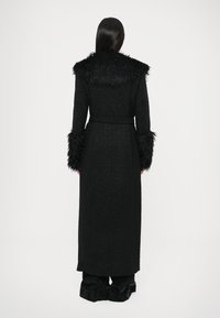 Dolgi črn plašč iz tkanine z teksturo, z ovratnikom in manšetami iz krzna. Vključuje usklajeni pas za prilagajanje v pasu.