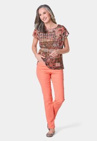Top a maniche corte con un motivo geometrico multicolore, abbinato a pantaloni corallo. Il modello indossa delle ballerine grigie, posando su uno sfondo bianco.