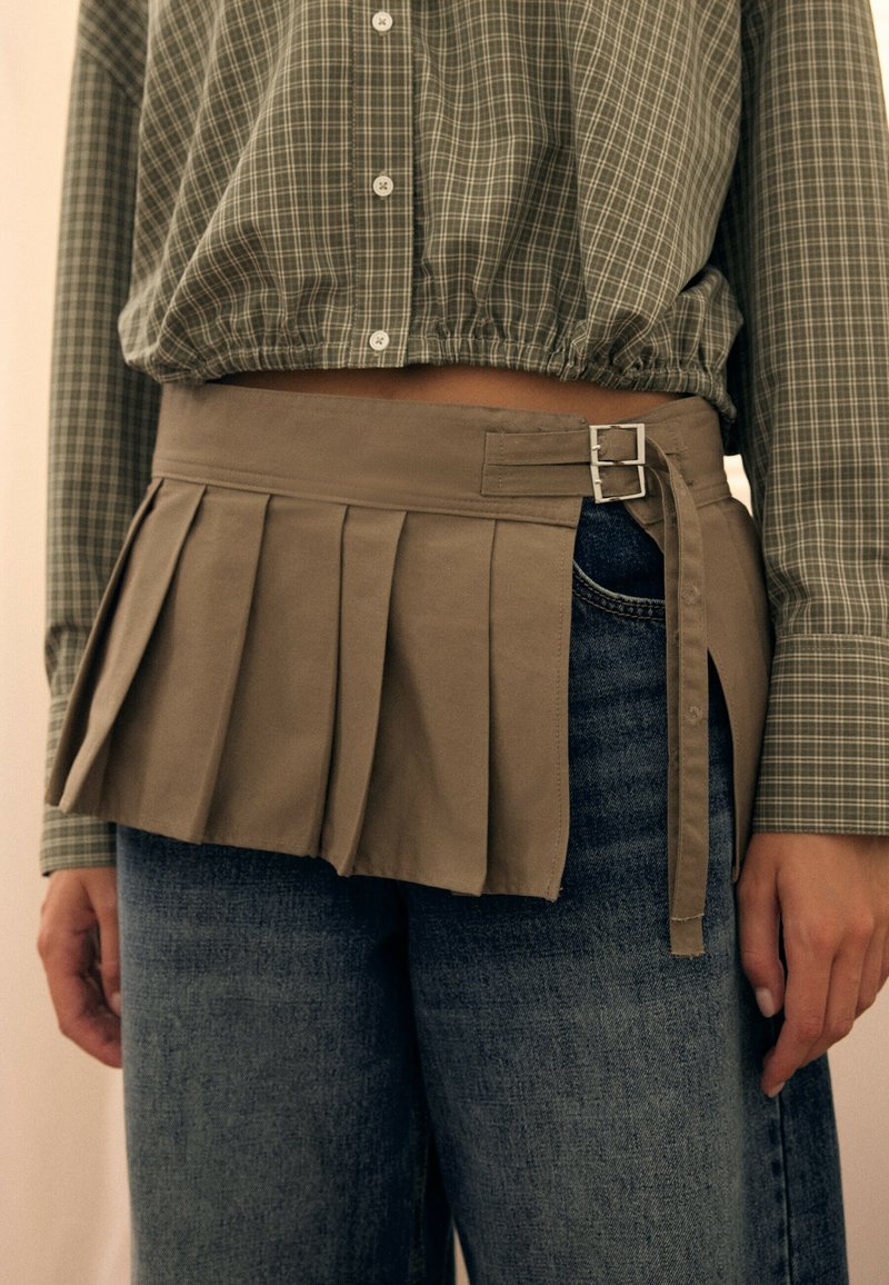 Personne portant une mini-jupe plissée marron avec ceinture sur un jean bleu et une chemise verte à carreaux boutonnée avec un ourlet élastique.