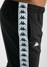 Pantalones deportivos negros con una franja lateral azul que presenta diseños en negro del logo repetidos. Cintura elástica y bolsillos laterales visibles.