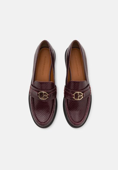 Mocasini din piele burgundy cu un finisaj texturat, având o cataramă aurie proeminentă și talpă din cauciuc negru. Design clasic, plat, fără șireturi.