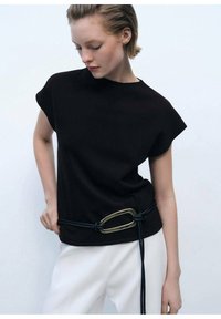 Black short-sleeve top with a round neckline, paired with white pants. A navy rope belt with a large gold buckle accents the waist.