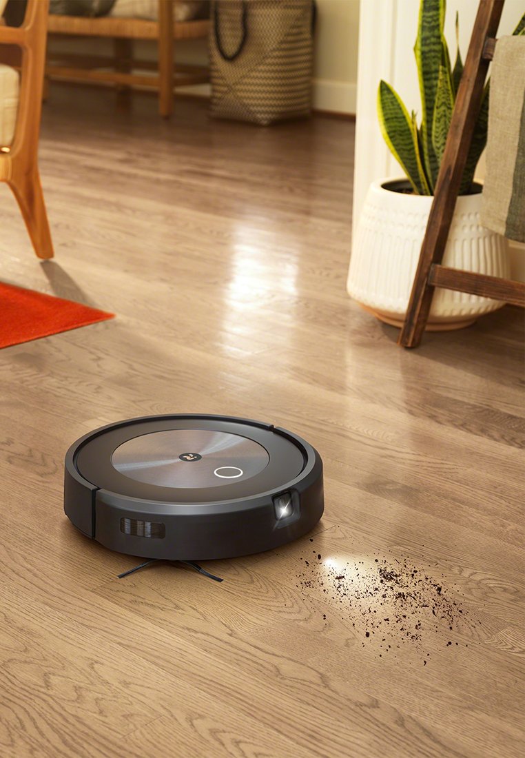 Ein schwarzer robotergestützter Staubsauger steht auf einem Holzboden, mit verstreutem Schmutz in der Nähe. Im Hintergrund sind ein Stuhl und eine Pflanze sichtbar.