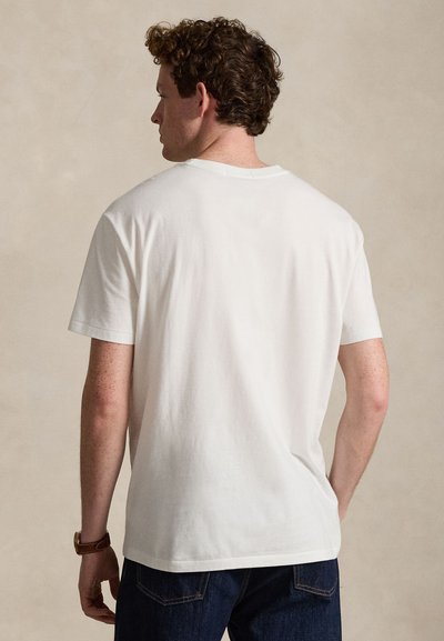 Mann mit lockigem Haar trägt ein schlichtes weißes kurzärmeliges T-Shirt und dunkelblaue Jeans, steht mit dem Rücken zur Kamera.