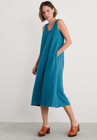 Teal cotton sleeveless dress with loose fit, knee-length hem, side pockets, and textured fabric. Paired with brown wooden platform clogs.