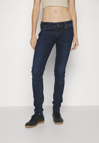 Jeans ajustados de color azul oscuro con una cintura de talle medio, bolsillos frontales y detalles de costura sutiles. Combinados con zapatillas negras sobre un fondo neutro.