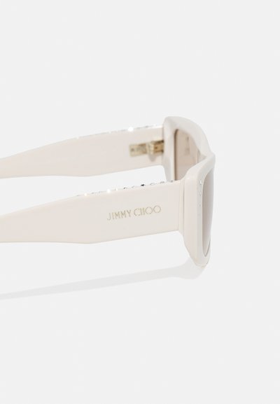 Las gafas de sol Jimmy Choo presentan un marco grueso de color crema, adornado con rhinestones a los lados y un sutil logo en dorado en la patilla.
