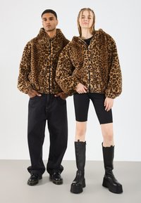 Bărbat și femeie stând în picioare, purtând jachete din blană artificială cu imprimeu leopard asortate; bărbatul are blugi negri și botine, iar femeia poartă pantaloni scurți negri și cizme cu talpă înaltă.