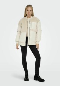 Giacca beige chiaro con una sezione superiore testurizzata e morbida e una cerniera frontale. Abbinata a pantaloni neri aderenti e stivali alla caviglia.