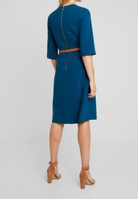 Tealfärgad klänning i knä längd med trekvartslånga ärmar och dragkedja i ryggen. Inkluderar ett smalt bälte i ljusbrunt och ett slätt, texturerat tyg.