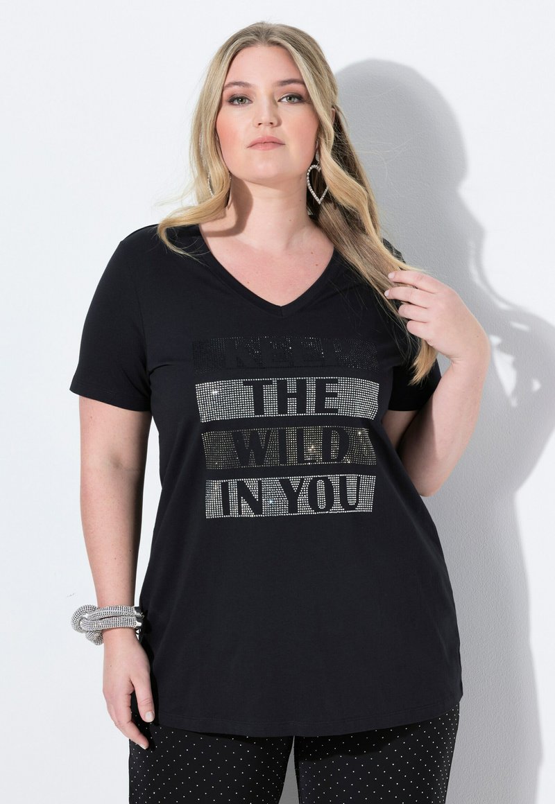 Černé tričko s véčkovým výstřihem, na kterém je text tvořený kameny říkající "UDRŽUJ DIVOKOST V SOBĚ." V kombinaci s černými kalhotami s puntíky.