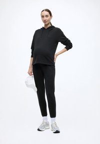 Czarna bluza z kapturem ze ściągaczem, czarne dopasowane spodnie, biała czapka w dłoni oraz szare buty sportowe. Gładka faktura, casualowy design.