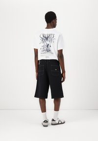 Vit t-shirt med palmgrafik och text på ryggen, kombinerad med svarta knälånga shorts. Bär vita strumpor och svart/vita sneakers.