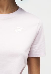 Άτομο που φοράει ανοιχτό ροζ T-shirt της Nike με λευκό λογότυπο Nike στο πάνω αριστερό μέρος του στήθους, μπροστά σε απλό ανοιχτό φόντο.